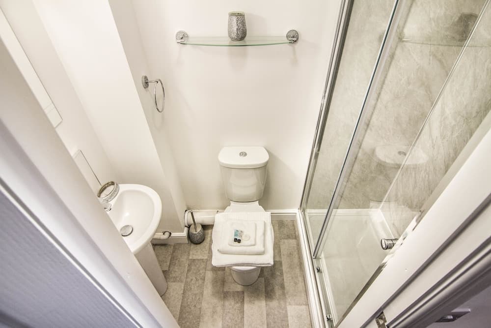 A cozy, compact bathroom features a sleek glass shower door, a pristine white toilet topped with folded towels, a small sink, and a minimalist shelf holding a decorative vase, ideal for travelers seeking efficient space utilization in accommodations.