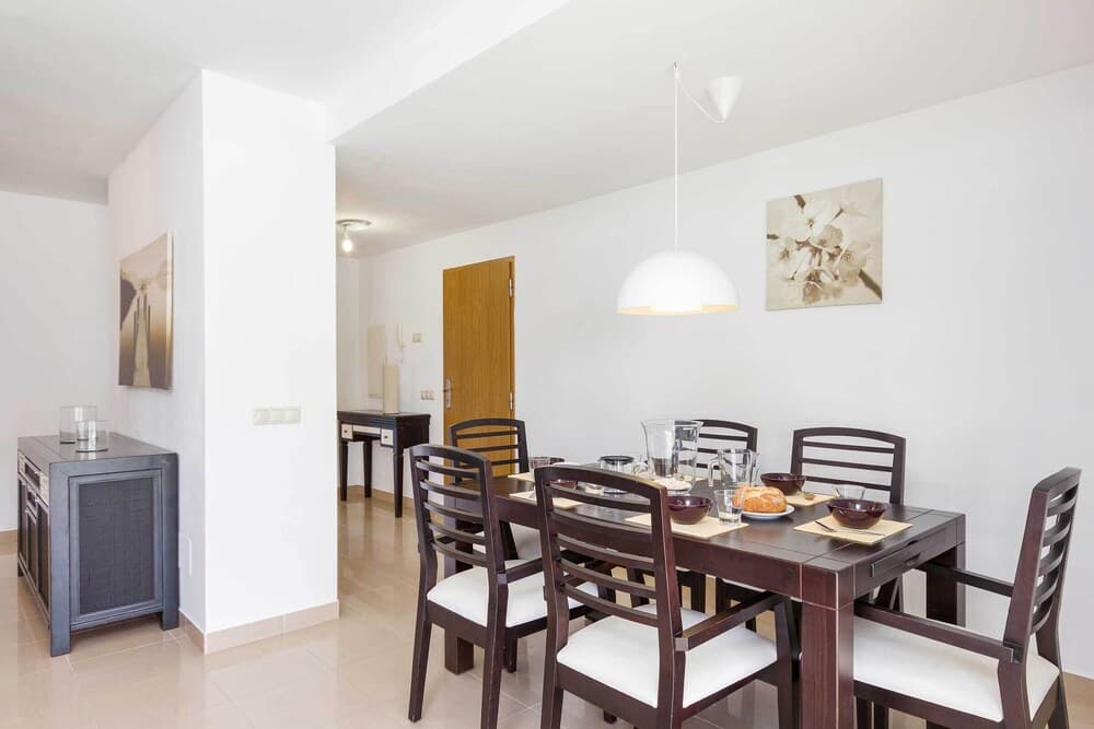 A cozy dining area with a wooden table set for breakfast, white cushioned chairs, modern pendant lighting, and minimalist decor, ideal for travelers seeking comfortable accommodations in a serene setting.