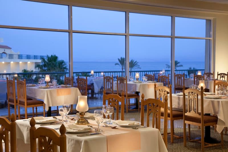 An elegant seaside restaurant with large windows overlooking the ocean at dusk, featuring warmly lit tables set for dining, ideal for romantic evenings or special occasions.