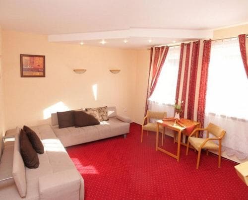 A cozy hotel room with beige sofas, red carpeting, wooden chairs, and curtains framing a bright window, ideal for travelers seeking comfort and relaxation.
