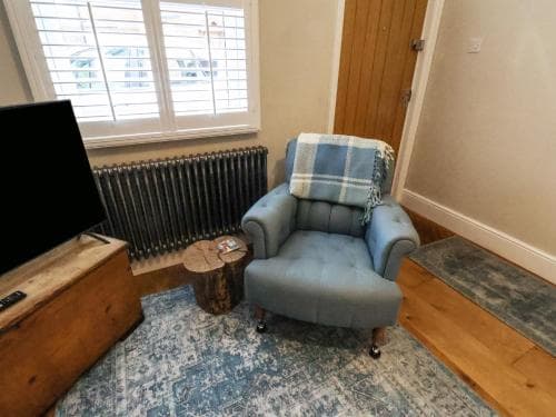 A cozy corner in a rustic living room features a plush blue armchair draped with a plaid blanket, set on a patterned rug beside a wooden TV stand and a small side table, ideal for travelers seeking comfort and relaxation.