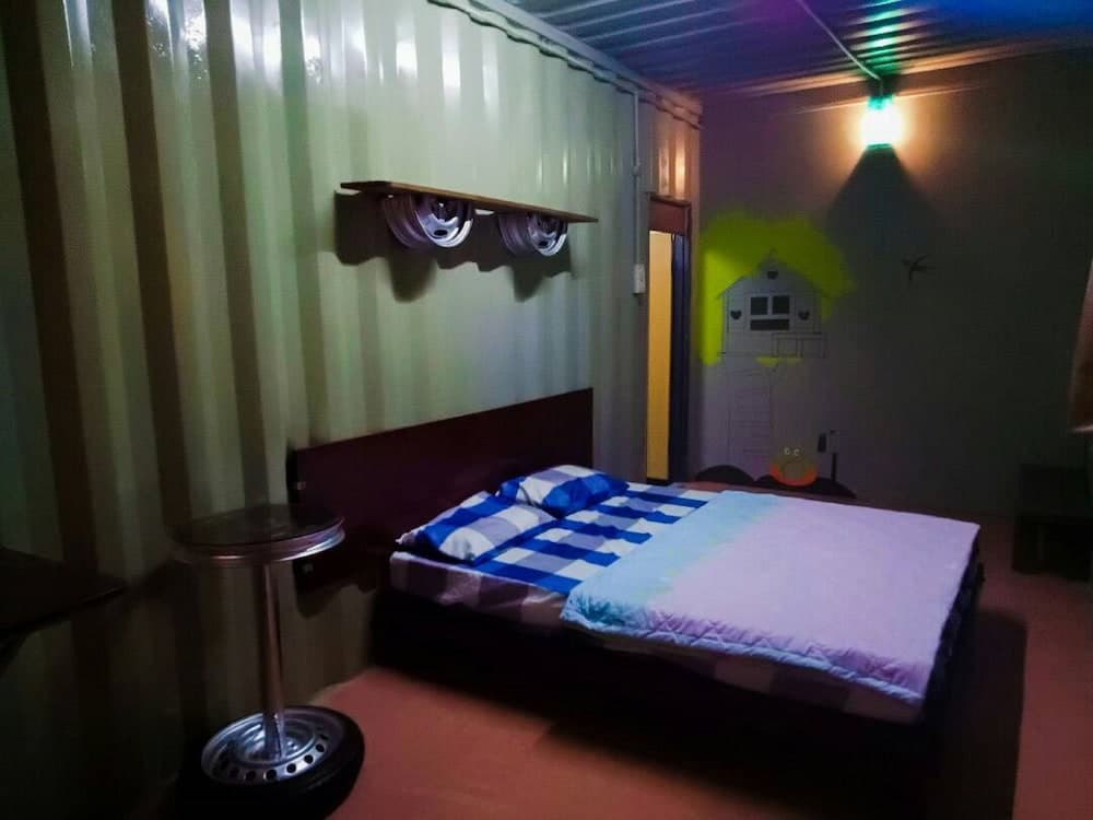 A cozy shipping container bedroom features a neatly made bed, quirky wall art, and repurposed wheel decor, creating an eclectic and inviting atmosphere ideal for budget travelers seeking unique accommodations.