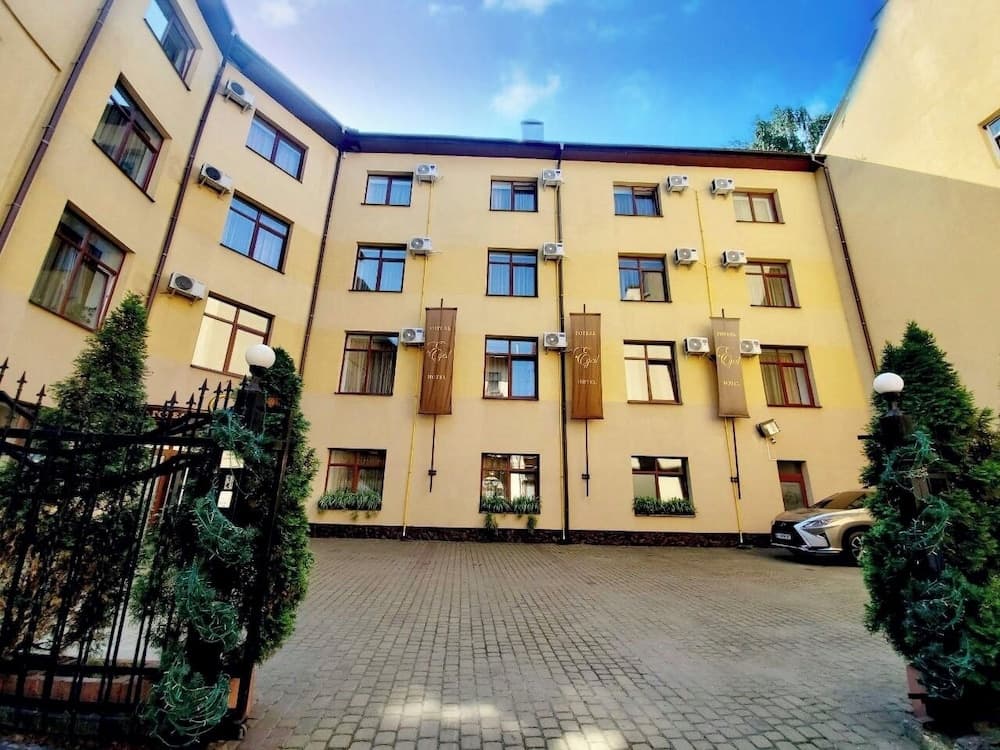 A serene courtyard with a beige hotel building adorned with air conditioning units and banners, framed by evergreen trees and a black wrought iron gate, ideal for travelers seeking a cozy urban retreat.