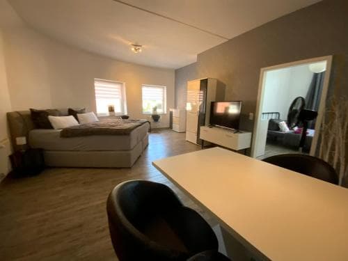 A cozy studio apartment features a neatly made bed by large windows, a sleek desk area, and an inviting living space, ideal for travelers seeking comfort and functionality in a modern urban retreat.