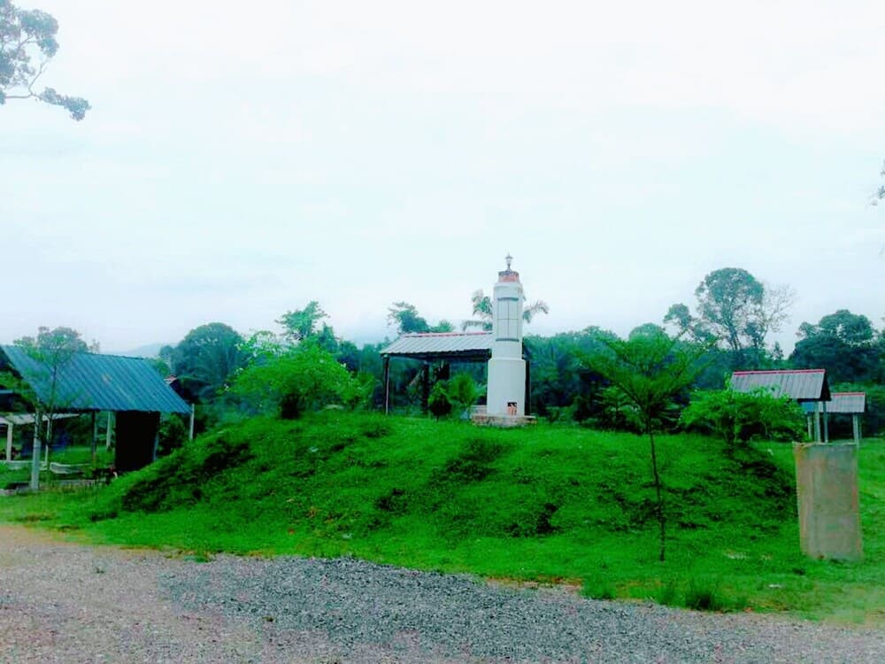 A serene rural landscape features a small white tower on a grassy mound surrounded by lush greenery and simple structures, ideal for tranquil countryside exploration.