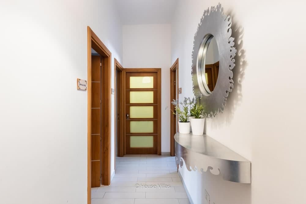 A modern villa hallway features sleek white walls, wooden doors, a decorative circular mirror, and a stylish console table with potted plants, ideal for showcasing interior design in vacation rental listings.
