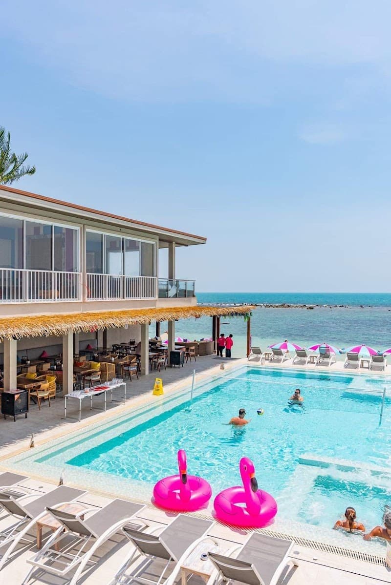 A vibrant beach club scene features a sparkling pool with pink flamingo floats, lounge chairs, and a thatched-roof dining area overlooking the turquoise sea, perfect for a relaxing tropical getaway.