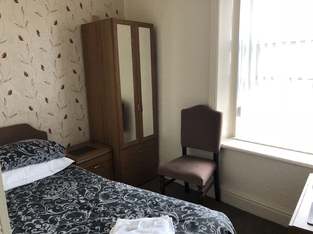 A cozy hotel room features a neatly made bed with patterned bedding, a wooden wardrobe with mirrored doors, a chair by the window, and floral wallpaper, ideal for travelers seeking comfort and simplicity.