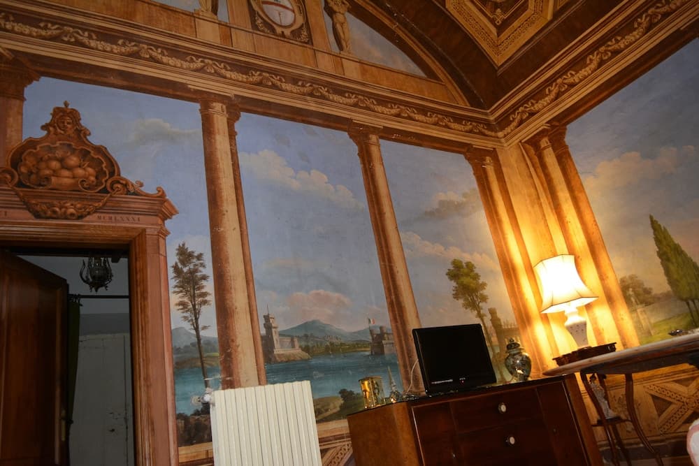 An opulent room features a stunning trompe-l'œil mural depicting a serene landscape with columns, enhancing the luxurious ambiance ideal for cultural tourism and historical exploration.