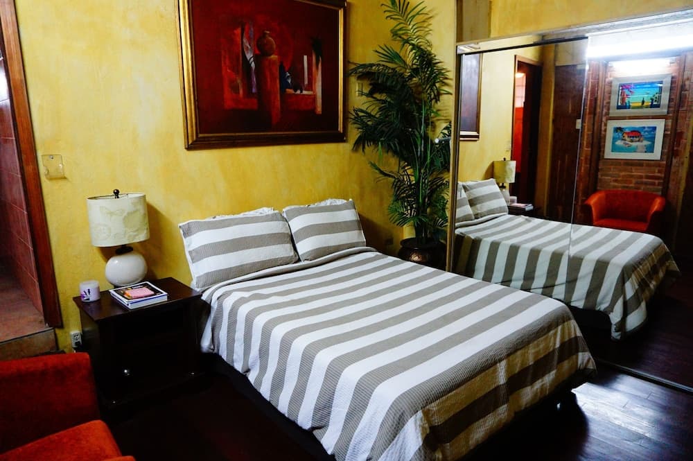 A cozy hotel room with warm yellow walls, striped bedding, a vibrant painting, and lush greenery creates an inviting atmosphere ideal for travelers seeking comfort and relaxation.