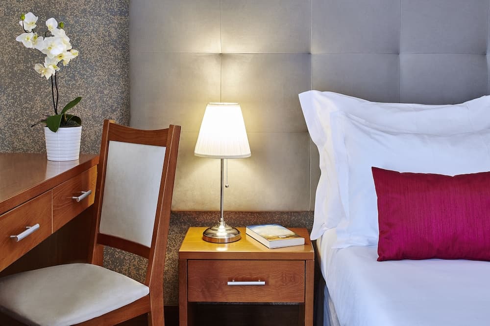 A cozy hotel room corner features a neatly made bed with white linens and a vibrant red pillow, a wooden desk with a chair, a glowing bedside lamp, and a vase of fresh orchids, ideal for travelers seeking comfort and relaxation.