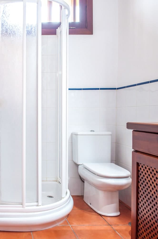 A cozy bathroom with white tiled walls, a corner shower enclosure, a toilet, and terracotta flooring offers a clean and functional space ideal for travelers seeking comfort in compact accommodations.