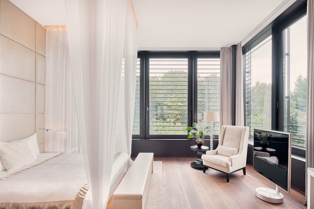 A serene modern bedroom with large windows overlooking lush greenery, featuring elegant furnishings like a plush bed, armchair, and minimalist decor, ideal for travelers seeking a tranquil retreat.