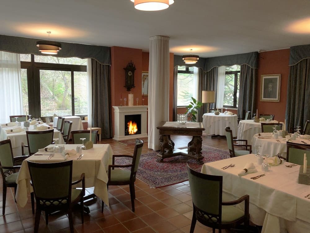 A cozy indoor dining area with warm lighting, a roaring fireplace, elegant tables set for guests, and large windows offering a serene view of greenery, ideal for a relaxing meal or event venue.