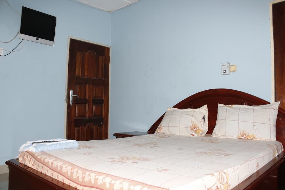 A cozy hotel room features a neatly made bed with floral bedding, a wooden door, a wall-mounted TV, and light blue walls, ideal for travelers seeking a simple and comfortable stay.