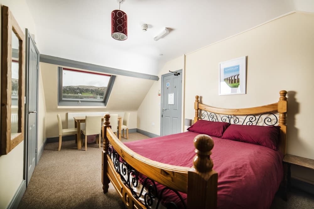 A cozy attic-style hotel room features a wooden bed with red bedding, a small dining area by the window overlooking scenic hills, and warm lighting, ideal for travelers seeking comfort and relaxation.