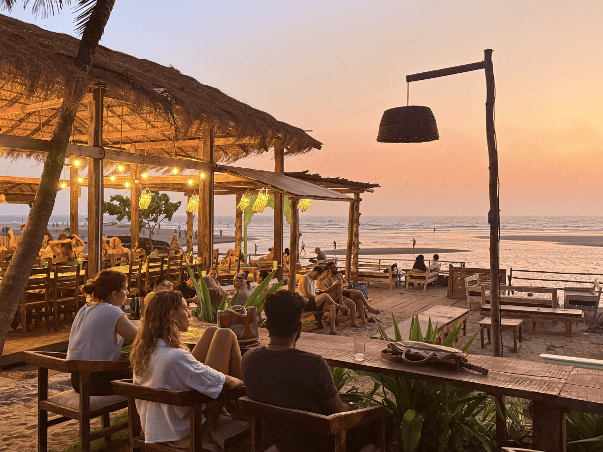 A serene beachside dining area at sunset, featuring rustic thatched roofs, twinkling lights, and patrons enjoying the tranquil ambiance, perfect for travelers seeking a relaxing coastal experience in Mandrem, Goa.