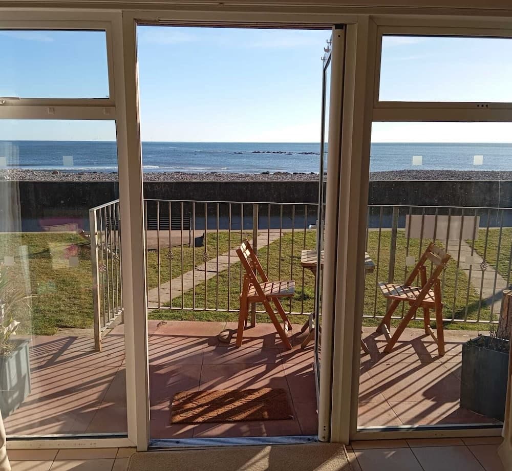 A serene coastal balcony view through open glass doors features wooden chairs, a calm sea, and a sunny atmosphere, ideal for relaxing seaside getaways.
