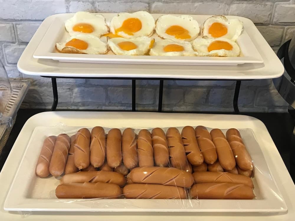 A cozy breakfast buffet scene featuring neatly arranged sunny-side-up eggs and hot dogs on tiered trays against a rustic brick wall backdrop, ideal for travelers seeking a hearty morning meal.