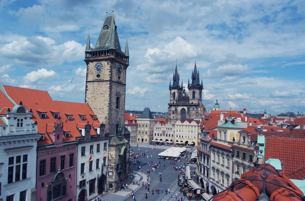 A vibrant European city square bustles under a partly cloudy sky, framed by historic architecture including a prominent clock tower and Gothic spires, ideal for cultural exploration and photography.