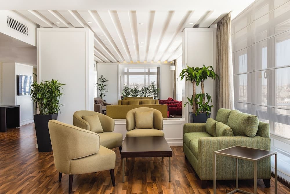 A cozy hotel lounge with plush green sofas, wooden flooring, indoor plants, and large windows offering city views, ideal for travelers seeking relaxation and comfort.