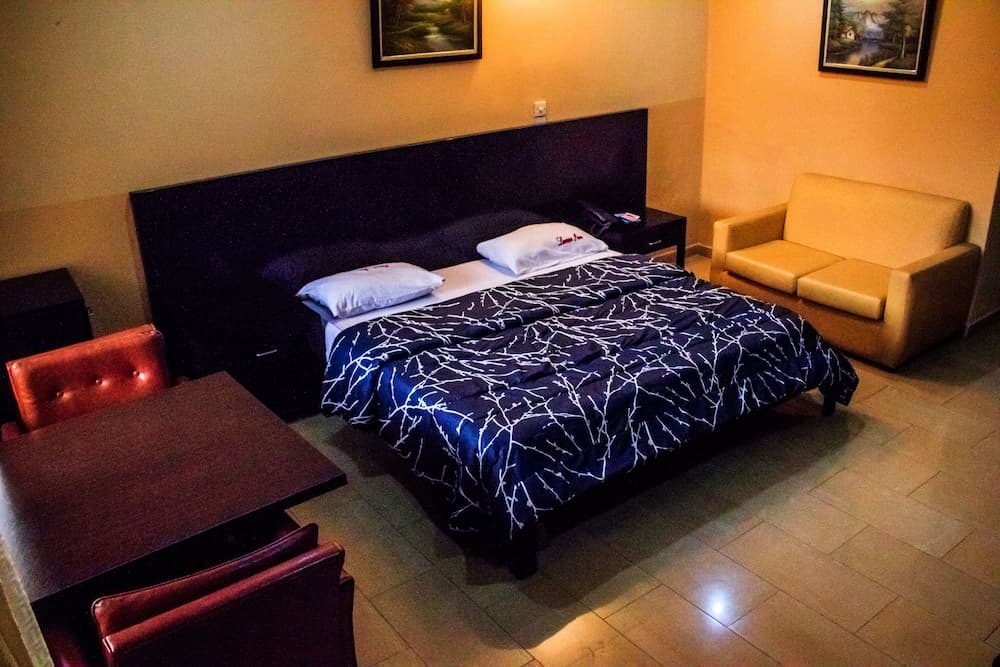 A cozy hotel room features a neatly made bed with a navy blue patterned duvet, a comfortable beige sofa, and a small dining table set, creating an inviting atmosphere ideal for travelers seeking relaxation.