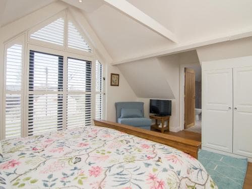 A cozy attic bedroom with sloped ceilings, large shuttered windows, and a floral bedspread creates a serene retreat, ideal for travelers seeking a charming and peaceful accommodation.