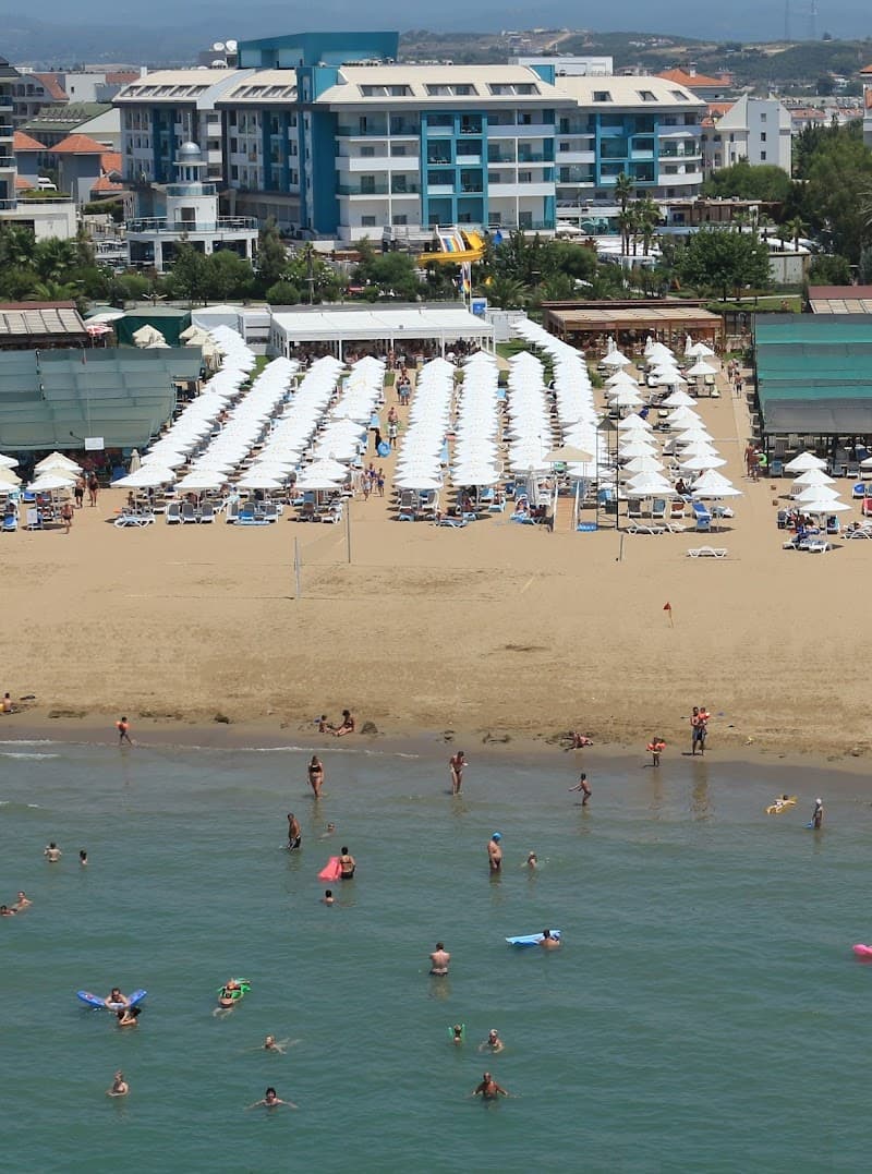 A vibrant beach scene at Seashell Resort & Spa in Antalya, Turkey, features rows of white umbrellas, sunbathers enjoying the sandy shore, and swimmers in the turquoise sea, creating a lively and relaxing atmosphere ideal for family vacations.