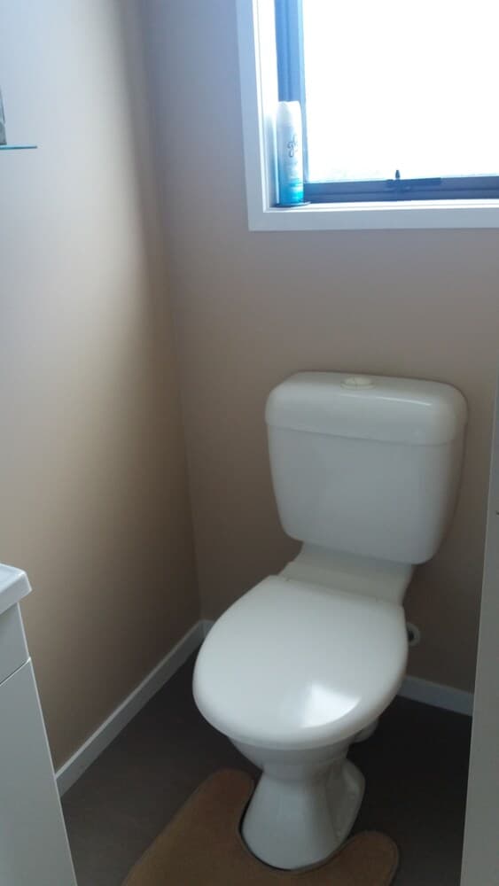 A minimalist bathroom corner features a white toilet beside a small window with a bottle of air freshener, creating a clean and functional space ideal for travelers seeking basic amenities.