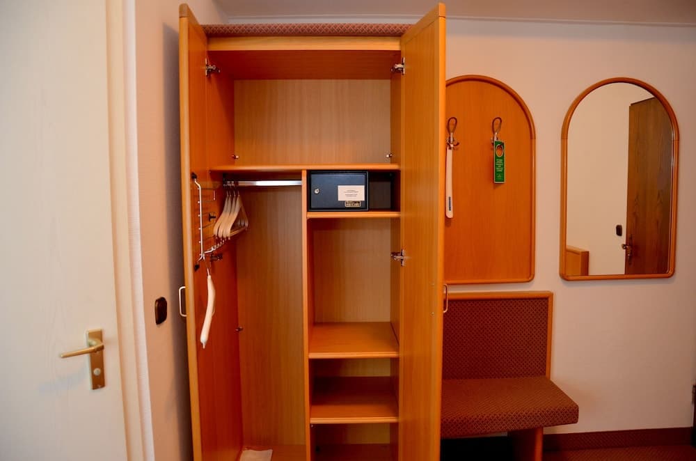 A cozy hotel room corner features an open wooden wardrobe with shelves, a safety deposit box, and hangers, complemented by a cushioned bench and an arched mirror, ideal for travelers seeking comfort and functionality.