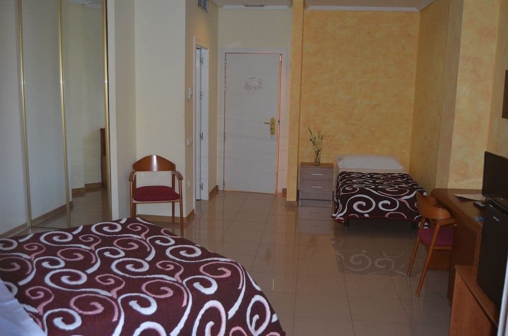A cozy hotel room features tiled floors, warm yellow walls, patterned beds, wooden furniture, and a serene ambiance ideal for travelers seeking comfort and simplicity.