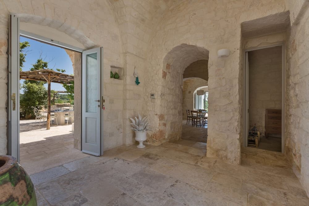 A serene stone-walled corridor with arched doorways leads to an outdoor patio, featuring rustic decor and natural light, ideal for travelers seeking a tranquil retreat in a historic setting.
