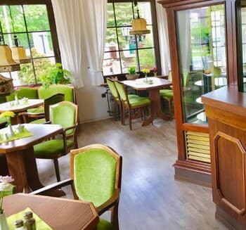 A cozy café with wooden tables, green chairs, and large windows inviting natural light creates a serene dining atmosphere, perfect for travelers seeking a relaxing meal spot.