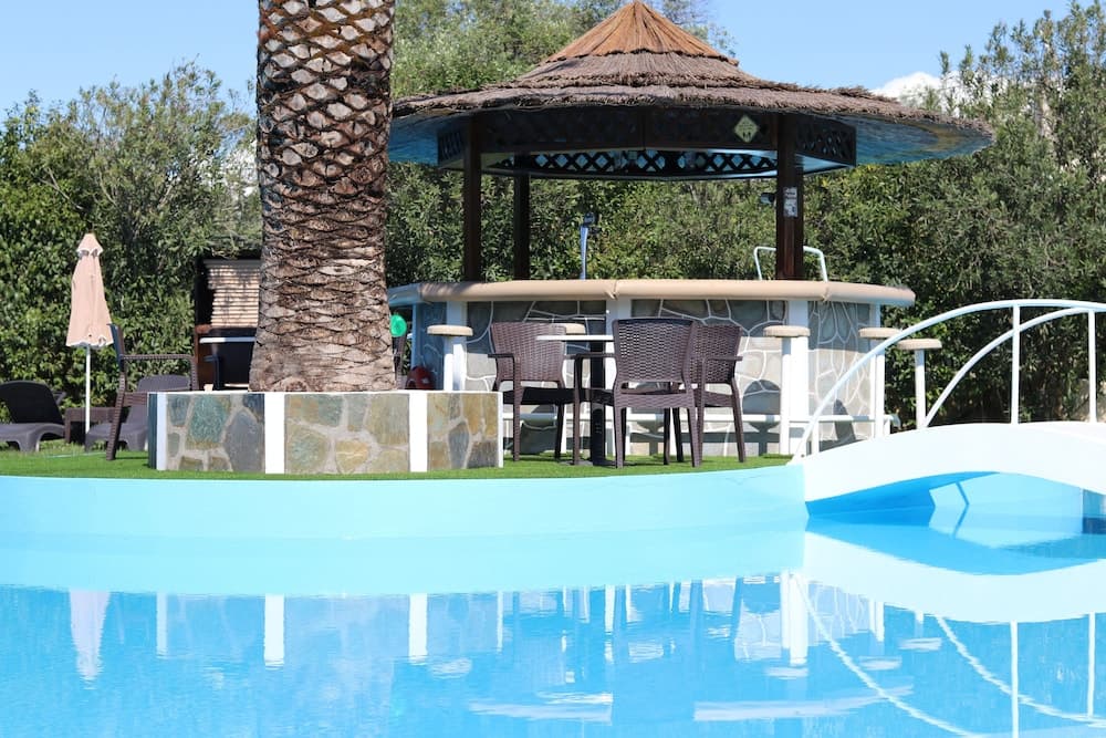 A serene resort poolside scene features a thatched-roof gazebo, palm tree, and lounge seating surrounded by lush greenery, ideal for relaxing vacations.