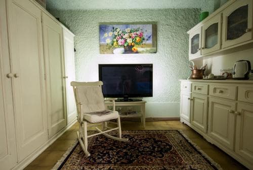 A cozy living room with a vintage aesthetic features a rocking chair on a patterned rug, a flat-screen TV, white cabinetry, and a floral painting, ideal for travelers seeking a homely retreat.