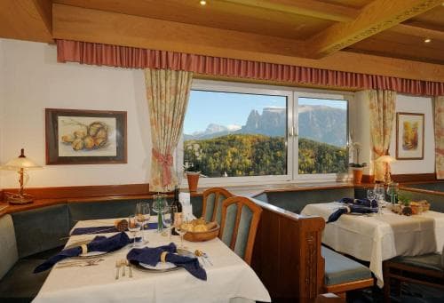 A cozy dining room with wooden accents and table settings offers a breathtaking view of lush mountains through large windows, ideal for travelers seeking a serene alpine retreat experience.