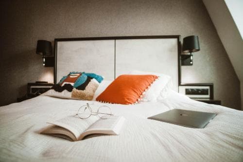 A cozy hotel room with a neatly made bed adorned with colorful pillows, an open book, glasses, and a laptop, evokes a serene and productive atmosphere ideal for travelers seeking relaxation and work-friendly accommodations.