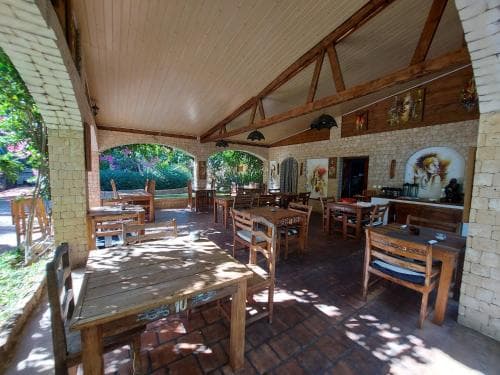 A cozy outdoor dining area with wooden tables and chairs under a shaded brick veranda, perfect for travelers seeking a relaxed meal in a rustic setting.