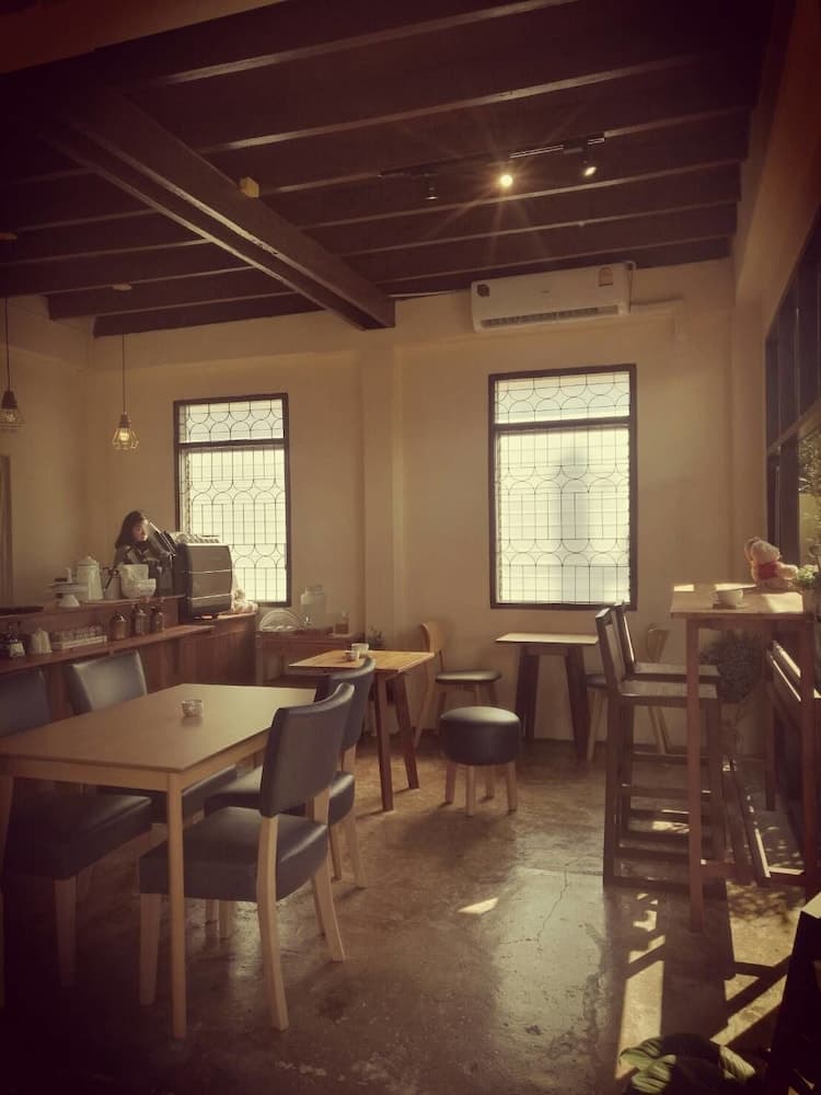 A cozy café with wooden tables, chairs, and exposed beams bathed in soft sunlight through patterned windows, ideal for travelers seeking a quiet spot to relax and enjoy a meal.