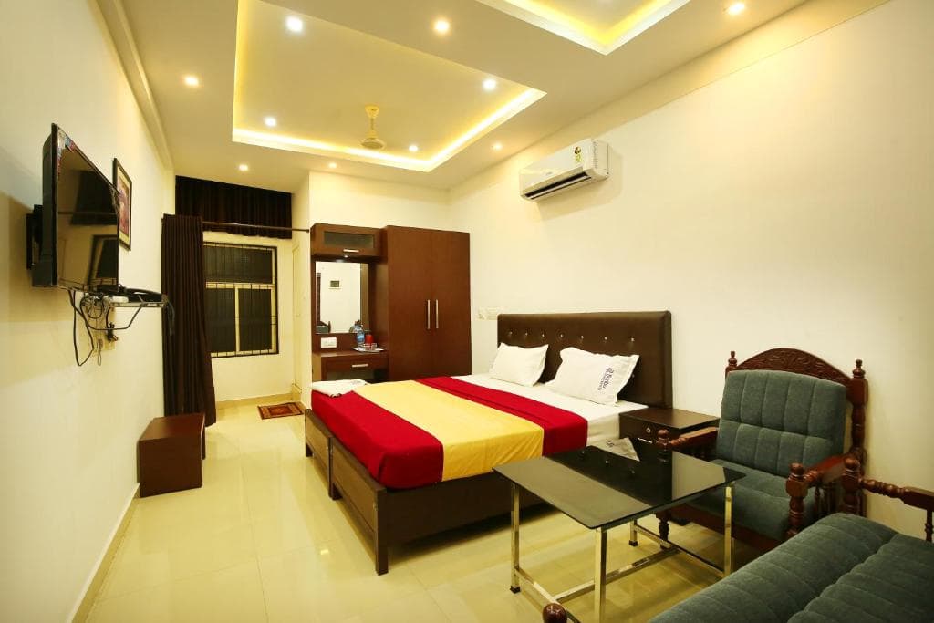 A cozy hotel room features a neatly made bed with red and yellow bedding, a green armchair, a flat-screen TV, and warm ambient lighting, ideal for travelers seeking comfort and relaxation.