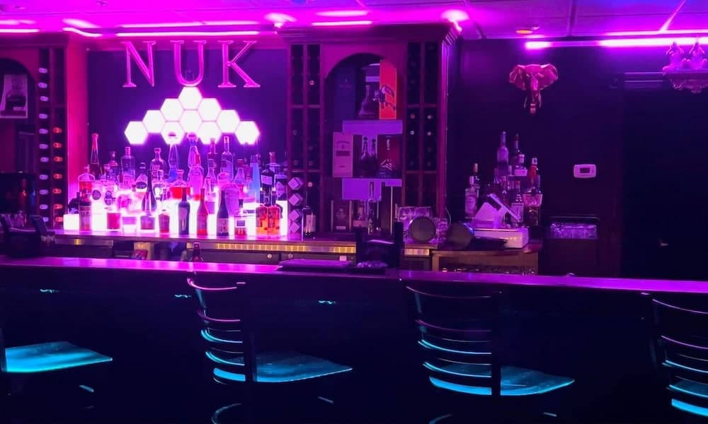 A vibrant nightclub bar glows under neon purple lights, featuring illuminated bottles, modern stools, and a striking "NUK" sign, ideal for nightlife enthusiasts seeking an energetic atmosphere.
