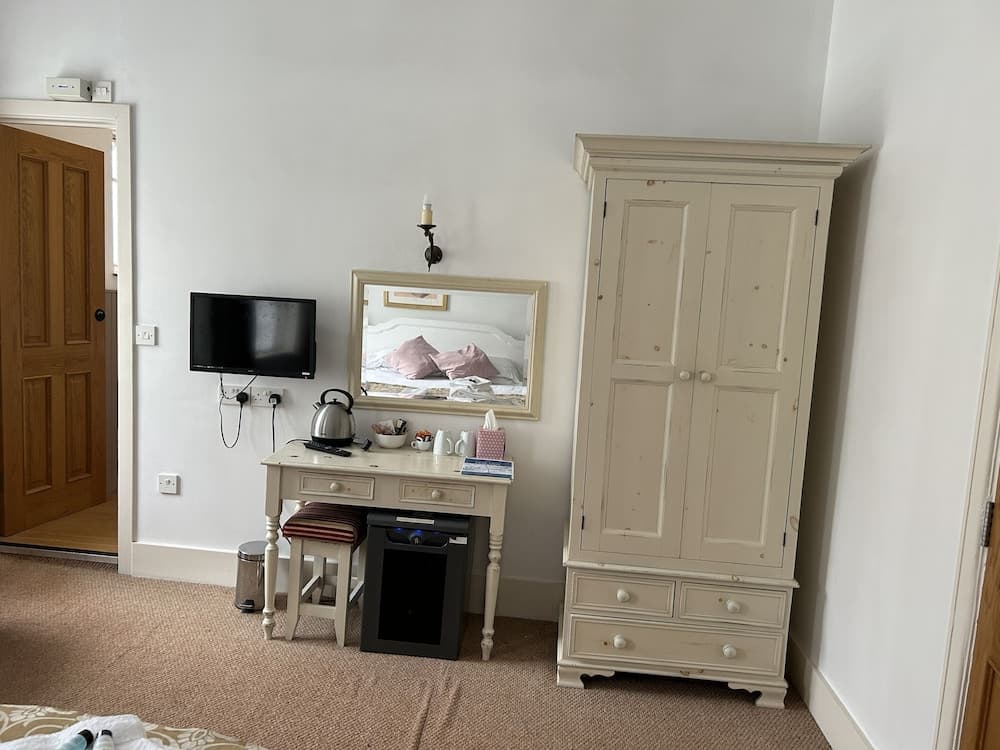 A cozy hotel room corner features a vintage-style dresser, a small desk with a kettle and mirror, and a mounted TV, creating a warm and inviting atmosphere ideal for travelers seeking comfort and charm.