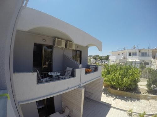 A serene Mediterranean-style apartment balcony with white walls, greenery, and clear blue skies offers a tranquil retreat for travelers seeking relaxation.