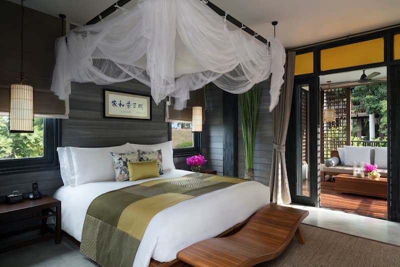 A serene tropical resort bedroom features a cozy bed under a flowing canopy, wooden furnishings, vibrant flowers, and a view of lush greenery through open doors, ideal for travelers seeking luxury and relaxation in Thailand.