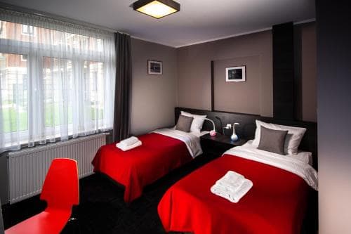 A cozy hotel room with two neatly made beds adorned in red linens, a vibrant red chair, and soft natural light filtering through sheer curtains, ideal for travelers seeking comfort and relaxation.