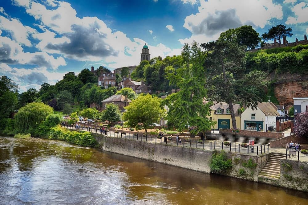 A picturesque riverside town with lush greenery, historic buildings, and a prominent church spire under a dramatic sky, ideal for tranquil walks and scenic photography.