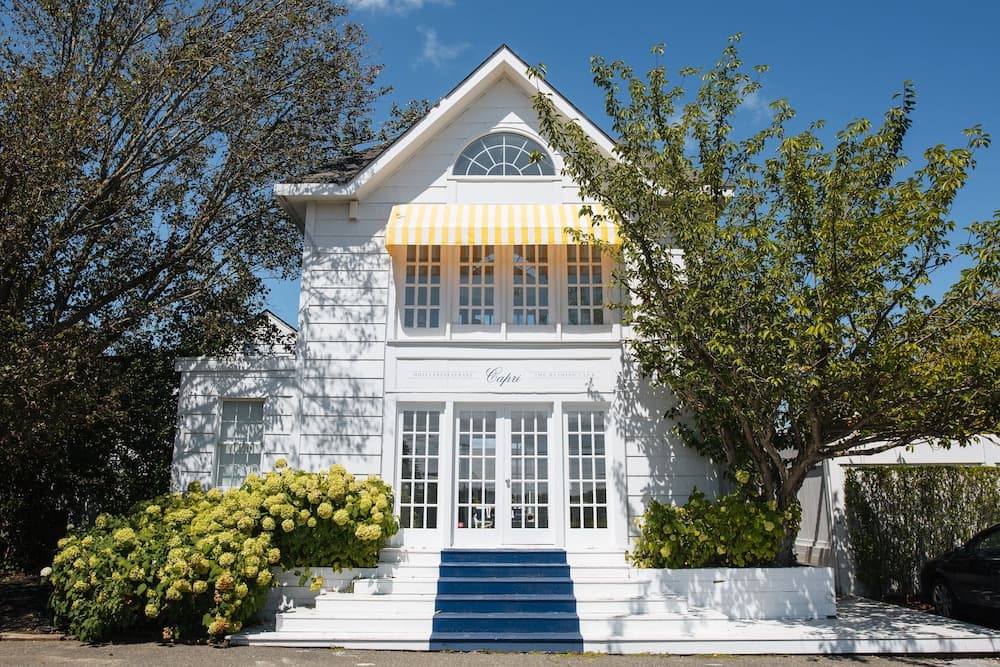 A charming white house with yellow-striped awnings and blue steps, surrounded by lush greenery under a clear blue sky, offers a picturesque setting ideal for a relaxing getaway or a scenic photo opportunity.
