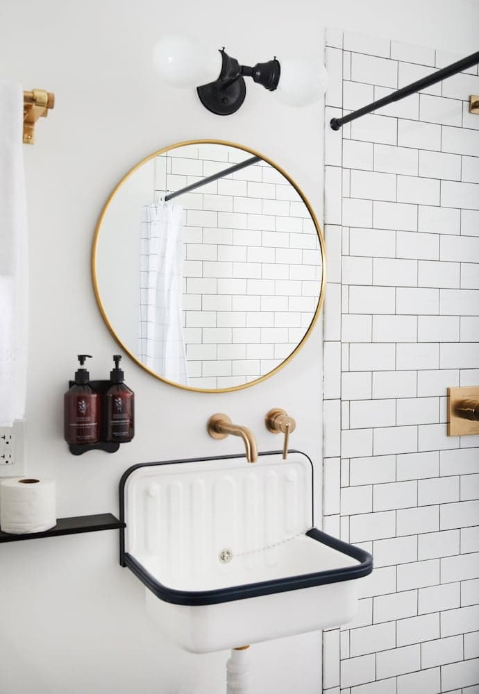 A modern bathroom features a sleek white sink with black trim, a round gold-framed mirror, and white subway tiles, creating a clean and stylish atmosphere ideal for travelers seeking contemporary accommodations.