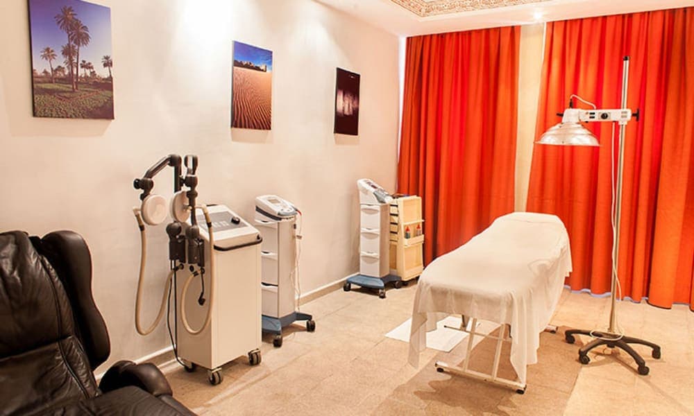 A serene spa treatment room features a massage table draped in white, vibrant orange curtains, medical equipment, and calming wall art, ideal for travelers seeking relaxation and wellness services.