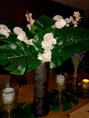 A cozy indoor dining setup features a centerpiece of white orchids in a black vase surrounded by lush green leaves, creating an elegant and serene atmosphere ideal for intimate gatherings or romantic dinners.